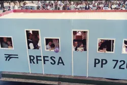 Uma fotografia colorida que registra um desfile ou evento público, onde um carro alegórico decorado como um vagão de trem da RFFSA (Rede Ferroviária Federal S.A.) transporta diversas pessoas, incluindo crianças. O cenário sugere uma celebração comunitária, possivelmente em uma cidade brasileira com tradição ferroviária, datada provavelmente da década de 1990 pelas vestimentas e qualidade da imagem.