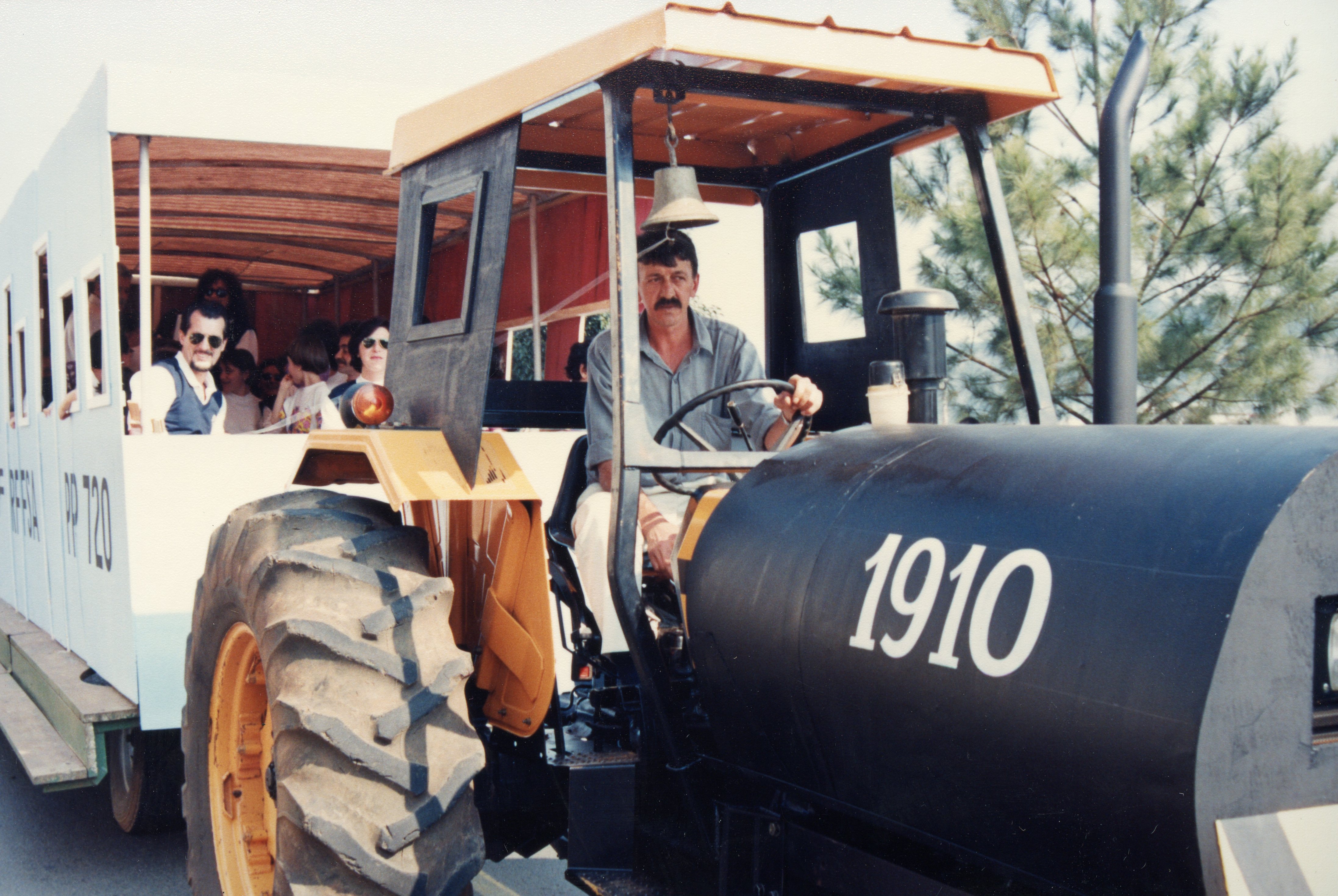 Uma fotografia colorida que mostra um trator adaptado para puxar um vagão de passageiros, possivelmente em um evento comemorativo ou turístico. O veículo ostenta o número 1910 e a sigla RFFSA, sugerindo uma homenagem à história ferroviária brasileira, com um motorista ao volante e passageiros visíveis no vagão.
