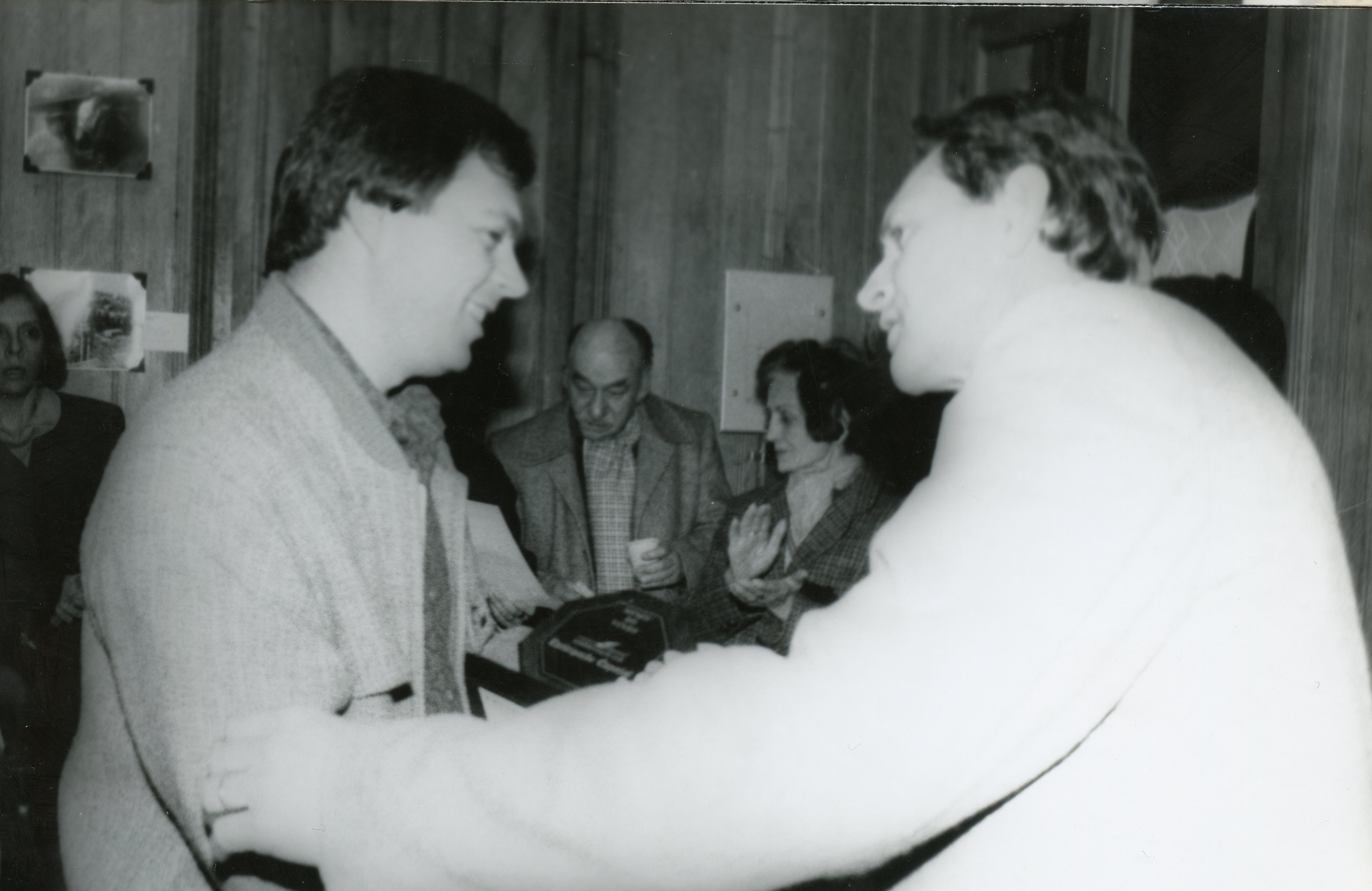 Fotografia em preto e branco registrando um momento de interação social ou premiação em um ambiente interno com paredes de madeira. O estilo das vestimentas e o corte de cabelo sugerem que a imagem foi capturada entre as décadas de 1980 e 1990, possivelmente em um evento comunitário ou corporativo.