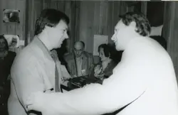 Fotografia em preto e branco registrando um momento de interação social ou premiação em um ambiente interno com paredes de madeira. O estilo das vestimentas e o corte de cabelo sugerem que a imagem foi capturada entre as décadas de 1980 e 1990, possivelmente em um evento comunitário ou corporativo.