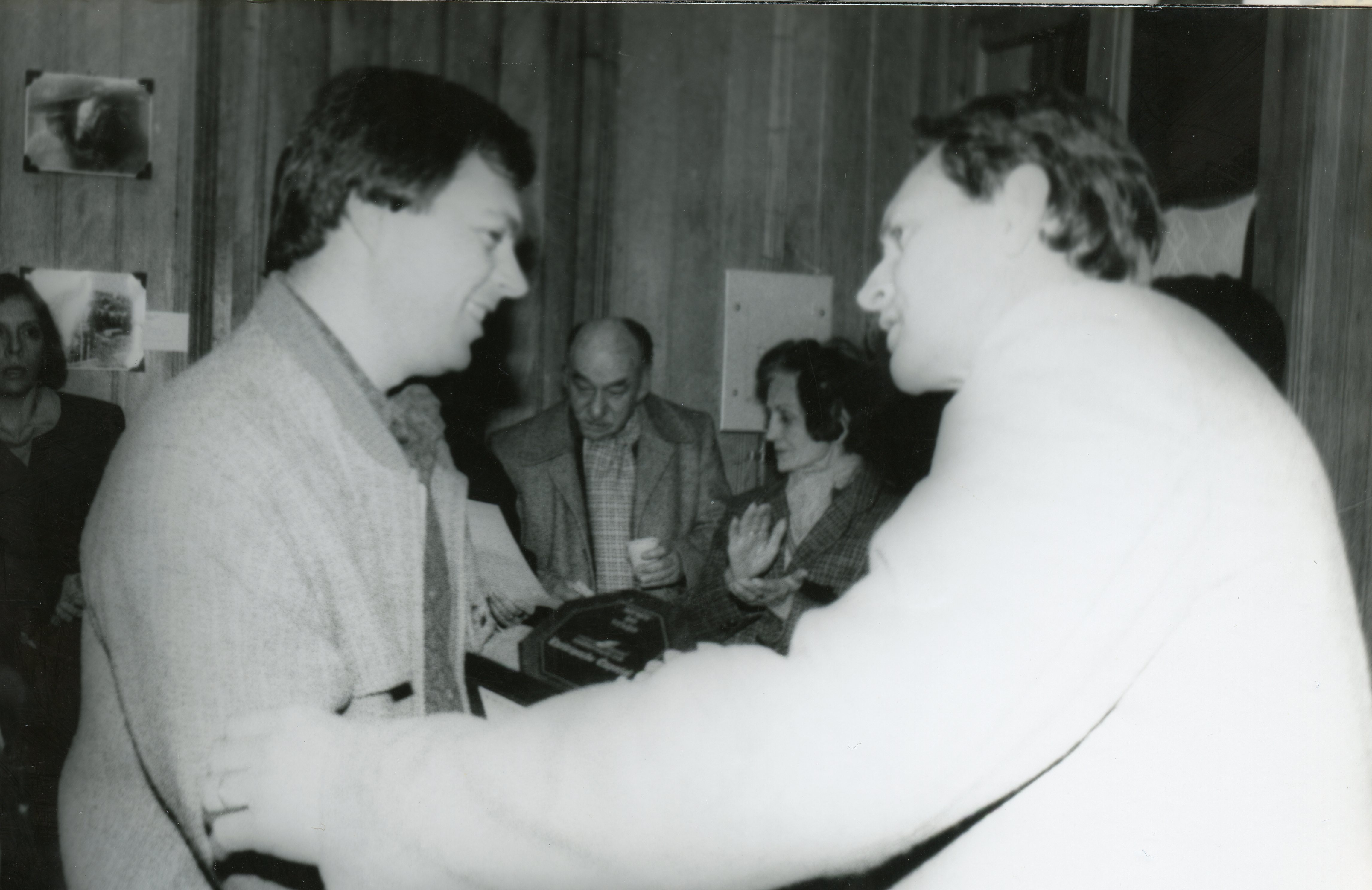 Uma fotografia em preto e branco que registra um momento de entrega de placa ou homenagem, possivelmente em um evento social ou corporativo. As pessoas vestem roupas formais típicas das décadas de 1980 ou 1990, em um ambiente interno com paredes de madeira.