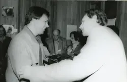 Uma fotografia em preto e branco que registra um momento de entrega de placa ou homenagem, possivelmente em um evento social ou corporativo. As pessoas vestem roupas formais típicas das décadas de 1980 ou 1990, em um ambiente interno com paredes de madeira.