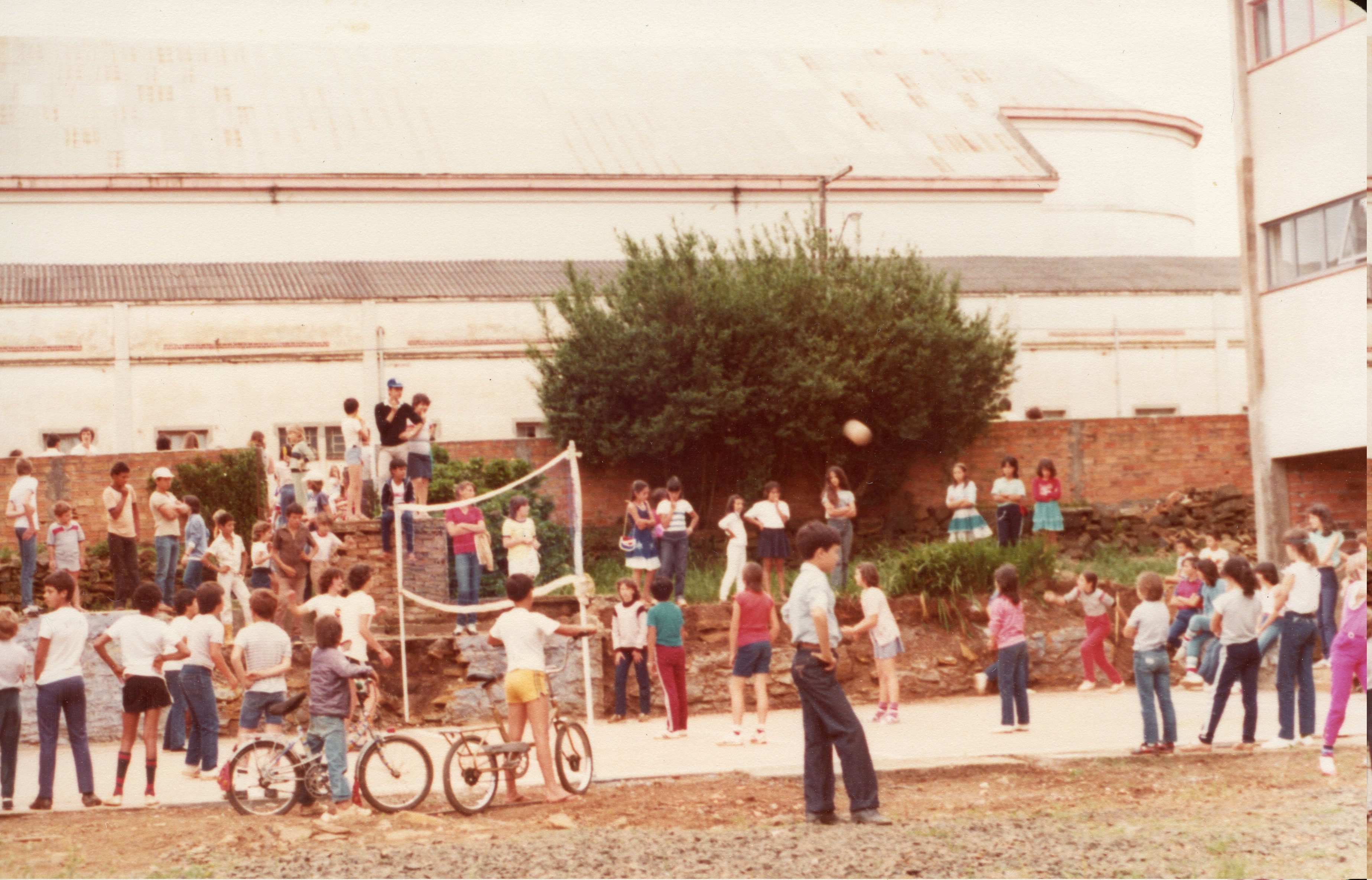 Uma fotografia colorida que retrata um grupo de crianças e adolescentes reunidos em uma quadra esportiva ao ar livre, possivelmente em um ambiente escolar ou recreativo. A cena apresenta vestimentas típicas das décadas de 1980 ou 1990, com a presença de bicicletas e uma rede de vôlei, sugerindo uma atividade de lazer comunitária em uma pequena cidade brasileira.