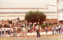 Uma fotografia colorida que retrata um grupo de crianças e adolescentes reunidos em uma quadra esportiva ao ar livre, possivelmente em um ambiente escolar ou recreativo. A cena apresenta vestimentas típicas das décadas de 1980 ou 1990, com a presença de bicicletas e uma rede de vôlei, sugerindo uma atividade de lazer comunitária em uma pequena cidade brasileira.