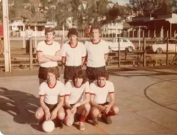 Uma fotografia de um time de futebol amador posando em uma quadra ao ar livre, provavelmente em uma cidade do interior brasileiro. Os jogadores vestem uniformes clássicos de camisas brancas com detalhes vermelhos e calções escuros, típicos da moda esportiva da década de 1970 ou início de 1980.
