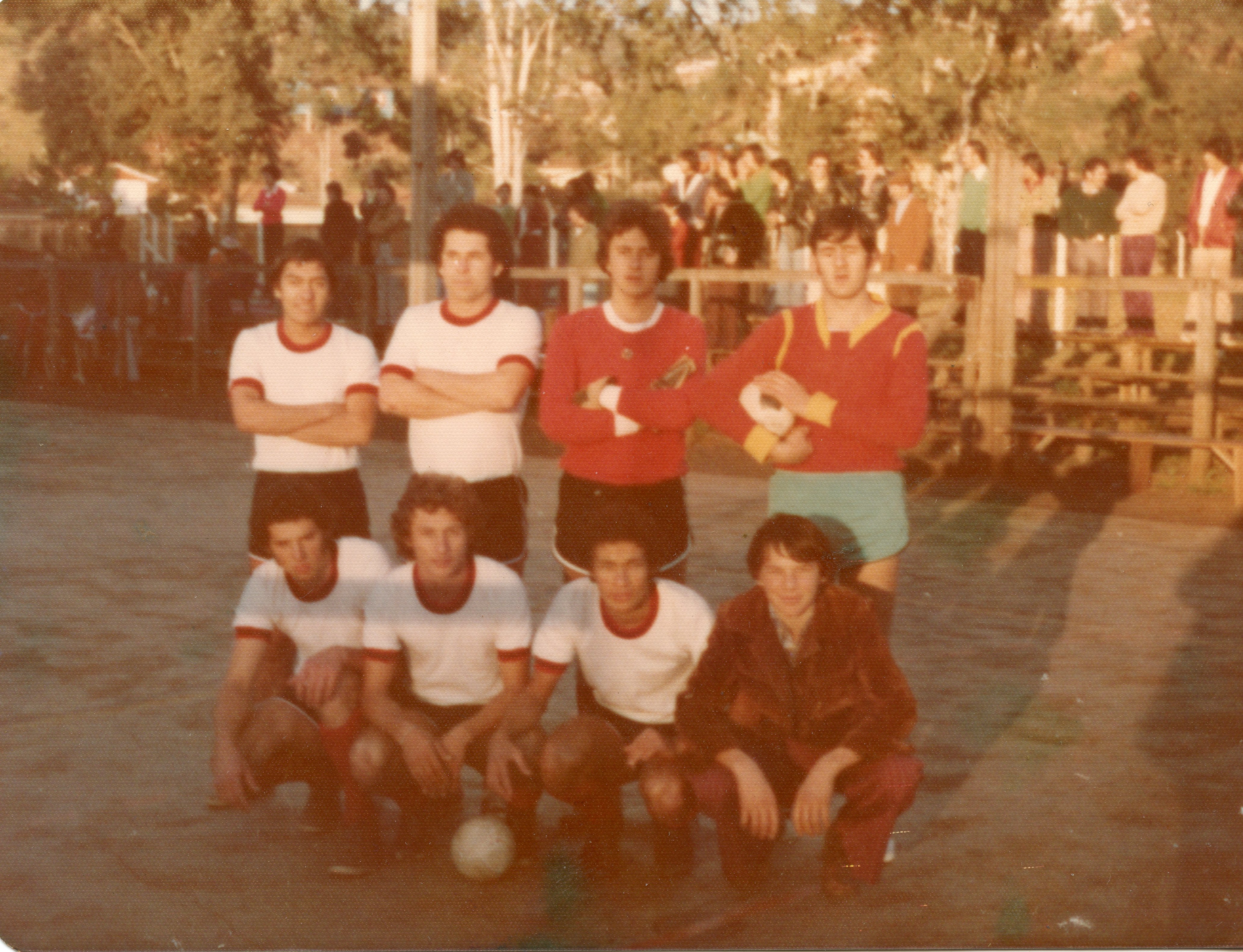 Uma fotografia de um time de futebol amador posando para uma foto em um campo de terra batida, provavelmente em uma pequena cidade brasileira. Os jogadores vestem uniformes típicos das décadas de 1970 ou 1980, com camisas de gola redonda e calções curtos, enquanto espectadores observam ao fundo atrás de uma cerca de madeira.