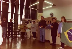 Uma fotografia de um evento cultural ou educacional em um ambiente interno, possivelmente uma biblioteca ou centro cultural, datada de maio de 1995. O grupo de pessoas está reunido, com uma jovem segurando a bandeira do Brasil, sugerindo uma cerimônia cívica ou abertura de atividades, como indicado pelo cartaz de 'Aulas de Flauta Doce'.