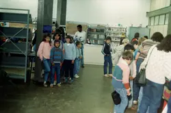 Uma cena interna que retrata um grupo de crianças e jovens em uma biblioteca ou centro cultural, provavelmente durante uma atividade escolar ou evento comunitário. A vestimenta e a qualidade da fotografia colorida sugerem que a imagem foi capturada entre o final da década de 1980 e início da década de 1990.