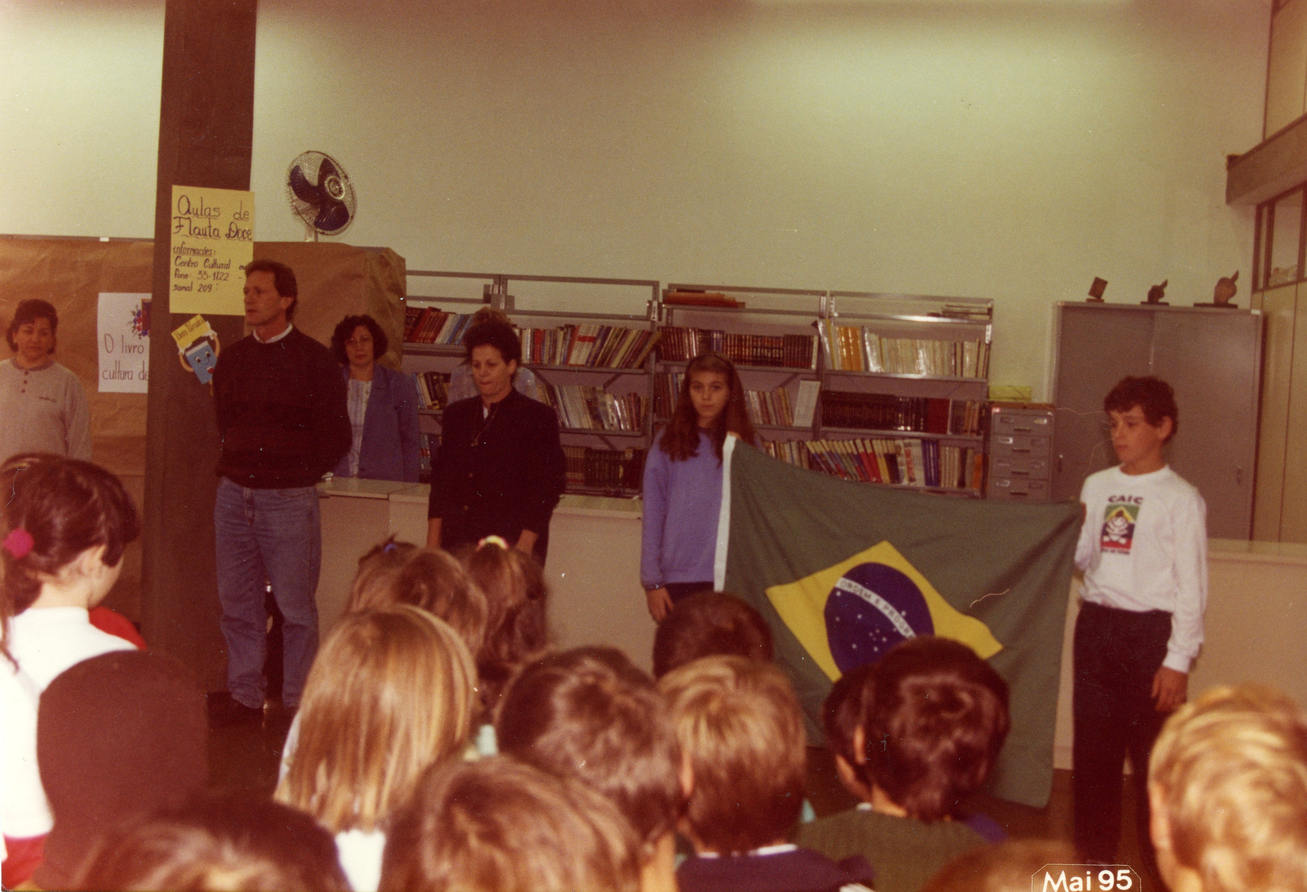 Uma cena escolar ou comunitária em um ambiente interno, possivelmente uma biblioteca, onde adultos e crianças estão reunidos. Um jovem segura a bandeira do Brasil enquanto outros observam, sugerindo uma cerimônia cívica ou evento educacional ocorrido em maio de 1995.