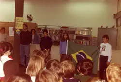 Uma cena escolar ou comunitária em um ambiente interno, possivelmente uma biblioteca, onde adultos e crianças estão reunidos. Um jovem segura a bandeira do Brasil enquanto outros observam, sugerindo uma cerimônia cívica ou evento educacional ocorrido em maio de 1995.