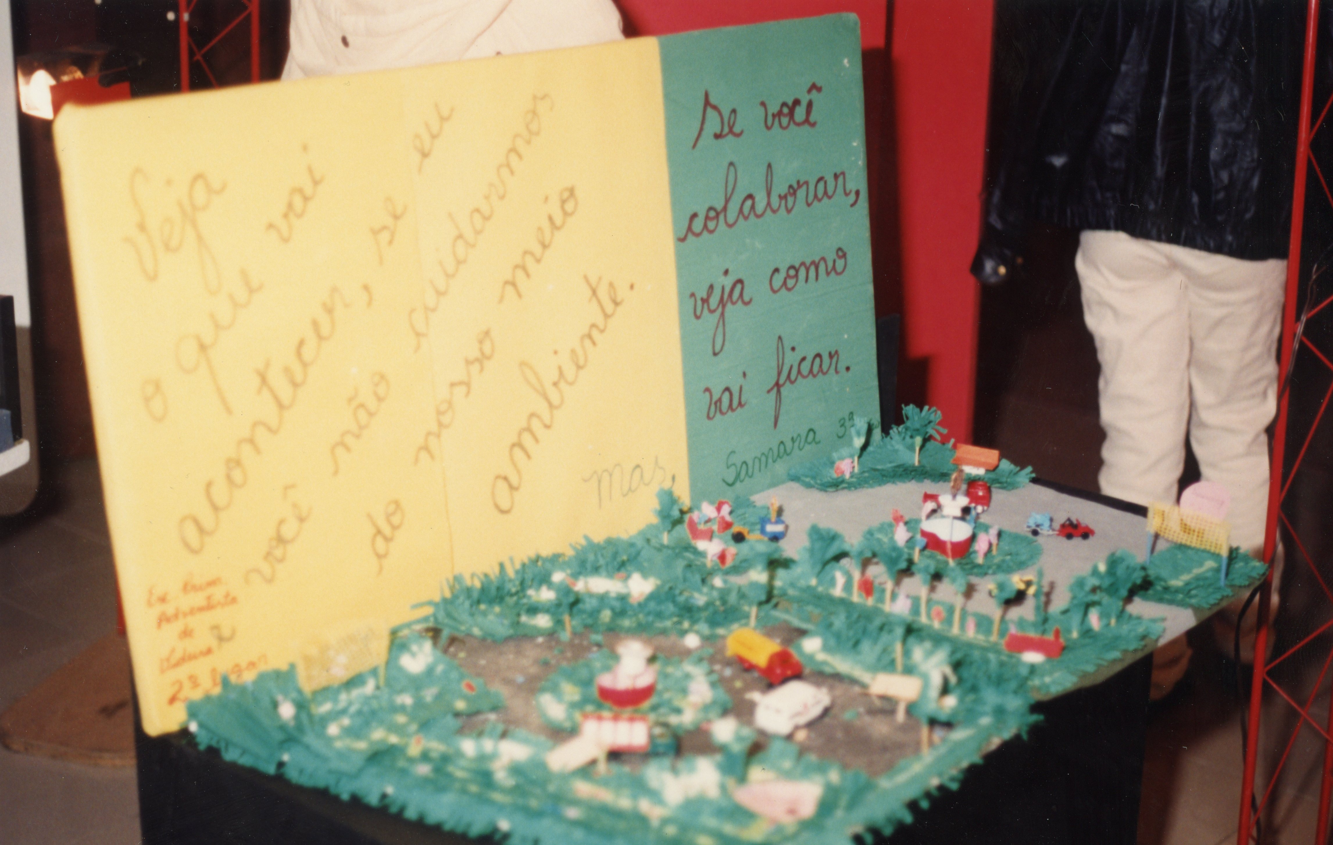Uma maquete escolar sobre preservação ambiental exibida em uma feira ou evento educacional. O cartaz, escrito à mão, traz uma mensagem reflexiva sobre o cuidado com o meio ambiente, possivelmente associado a uma escola em Videira, dado o contexto da assinatura.