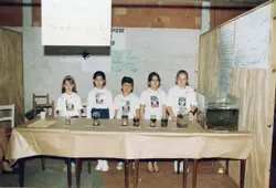 Uma fotografia de cinco crianças em uma feira de ciências escolar, provavelmente em um projeto CAIC, apresentando um estudo sobre a metamorfose dos anfíbios. Elas estão atrás de uma mesa com potes de vidro contendo espécimes e cartazes educativos, vestindo camisetas uniformizadas com o logo do programa.