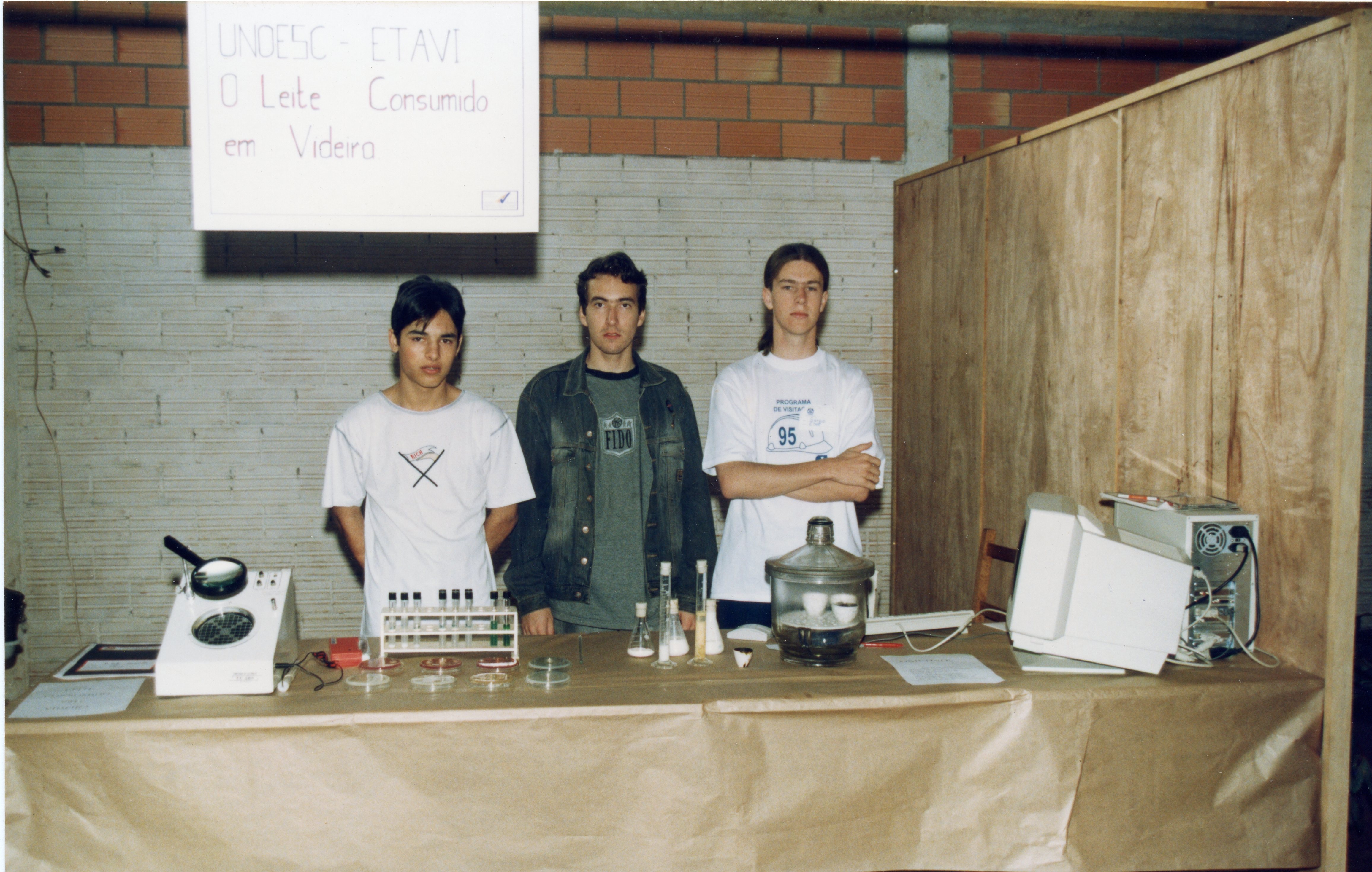 Três jovens posam atrás de uma bancada em uma feira de ciências ou evento acadêmico em Videira, apresentando um projeto sobre a qualidade do leite. A cena, datada de meados da década de 1990, exibe equipamentos laboratoriais, um computador antigo e cartazes informativos em um ambiente com paredes de tijolos aparentes.