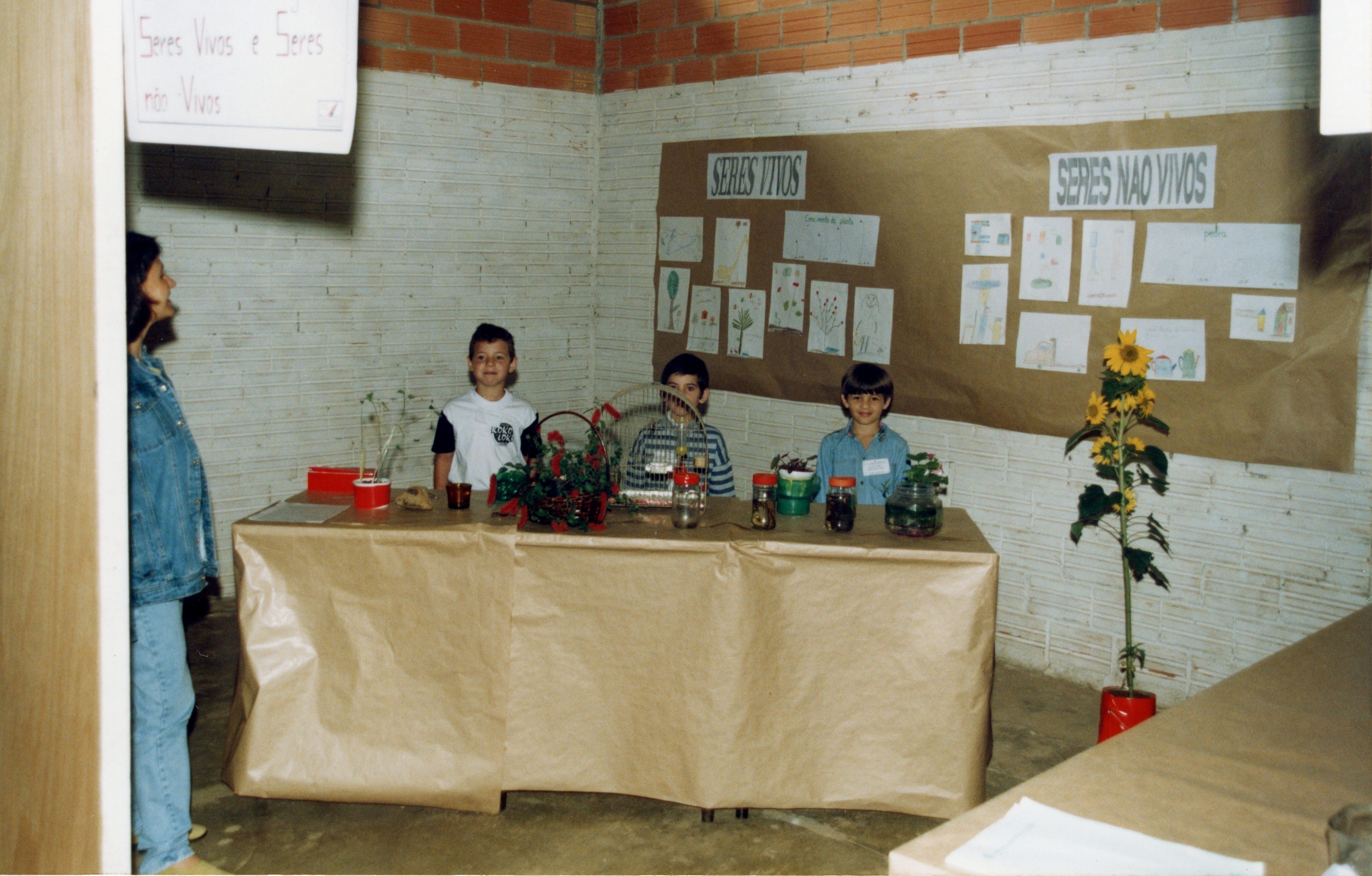 Uma fotografia de uma feira de ciências escolar, provavelmente realizada no final da década de 1990 ou início dos anos 2000. Três crianças estão atrás de uma mesa coberta com papel pardo, exibindo um projeto sobre 'seres vivos e seres não vivos', com desenhos e plantas, enquanto uma adulta observa ao lado.