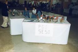 Uma feira escolar ou evento educacional com mesas cobertas por toalhas brancas, exibindo potes de vidro com amostras biológicas. O ambiente parece ser um ginásio ou salão comunitário, com jovens e adultos circulando, sugerindo uma atividade pedagógica do SESI possivelmente na década de 1990.