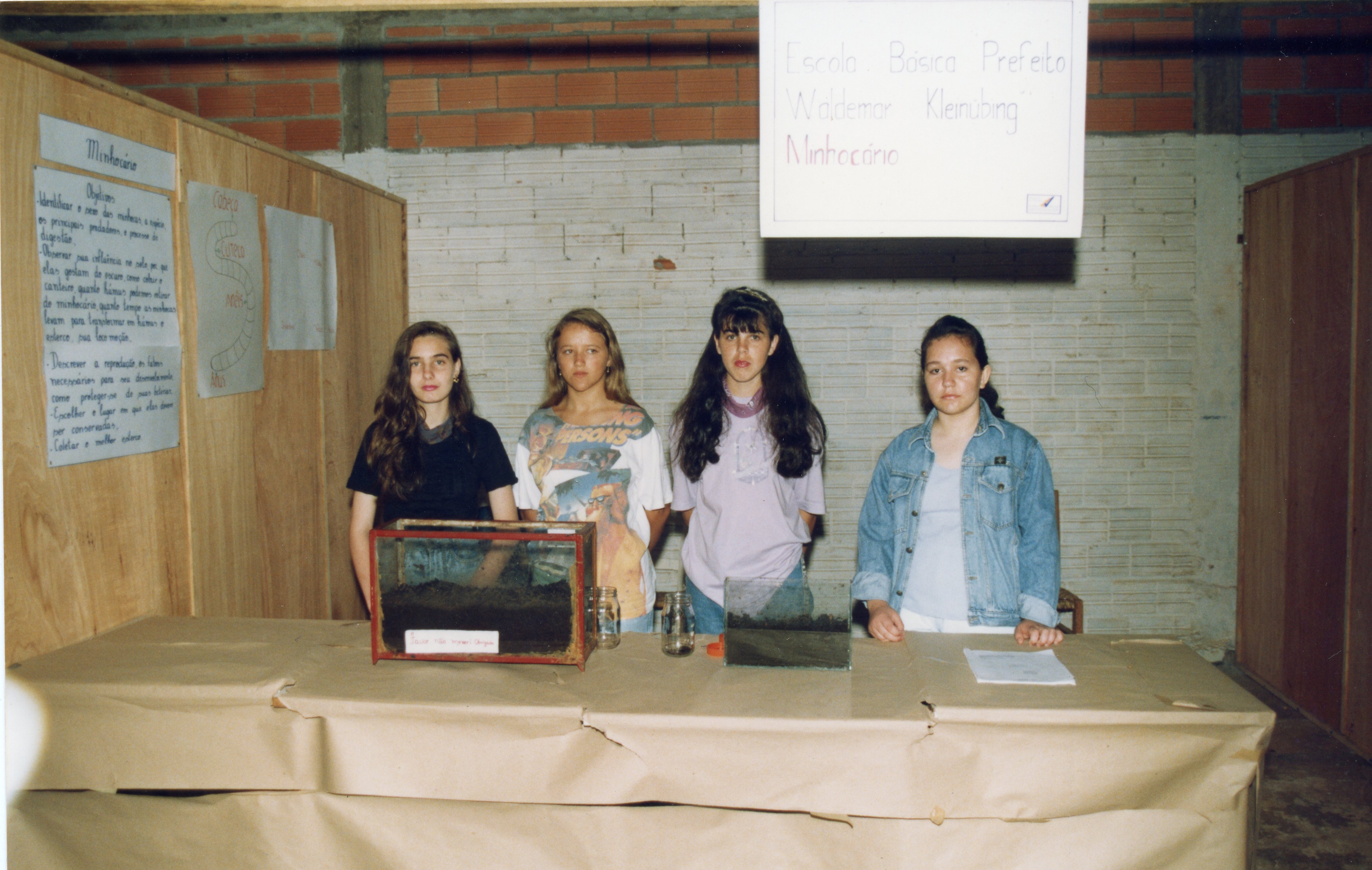Quatro estudantes posam atrás de uma mesa em uma feira de ciências escolar, apresentando um projeto sobre minhocário. O ambiente é uma sala com paredes de tijolos aparentes, típica de instituições de ensino brasileiras da década de 1990, com cartazes informativos e recipientes de vidro contendo terra.