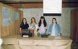 Quatro estudantes posam atrás de uma mesa em uma feira de ciências escolar, apresentando um projeto sobre minhocário. O ambiente é uma sala com paredes de tijolos aparentes, típica de instituições de ensino brasileiras da década de 1990, com cartazes informativos e recipientes de vidro contendo terra.