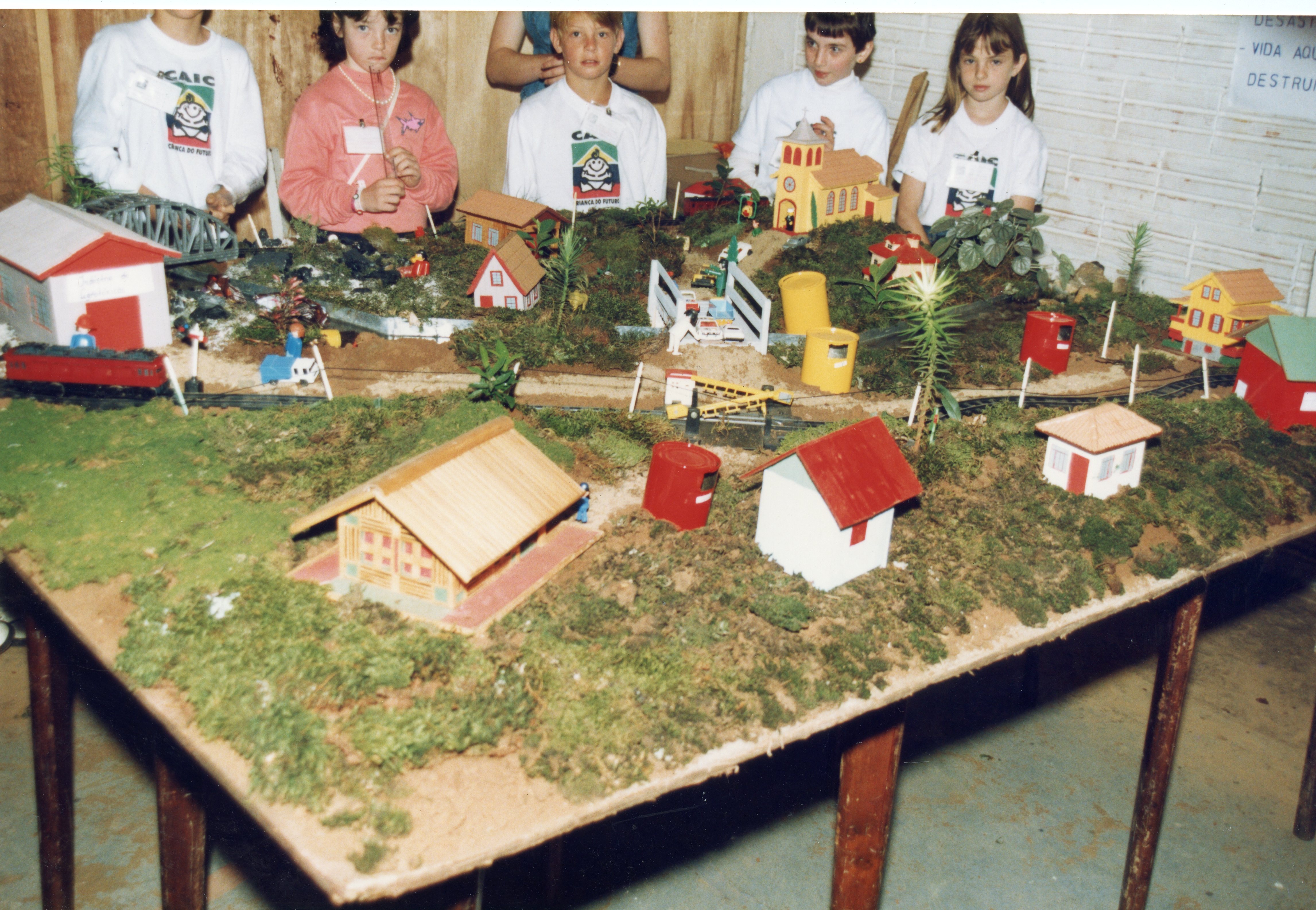 Uma fotografia colorida mostrando um grupo de crianças em uma feira escolar ou evento educacional, apresentando uma maquete detalhada de uma pequena cidade ou vila. As crianças vestem camisetas do projeto CAIC, sugerindo um contexto escolar brasileiro da década de 1990, com elementos como ferrovias, casas em miniatura e vegetação artificial.