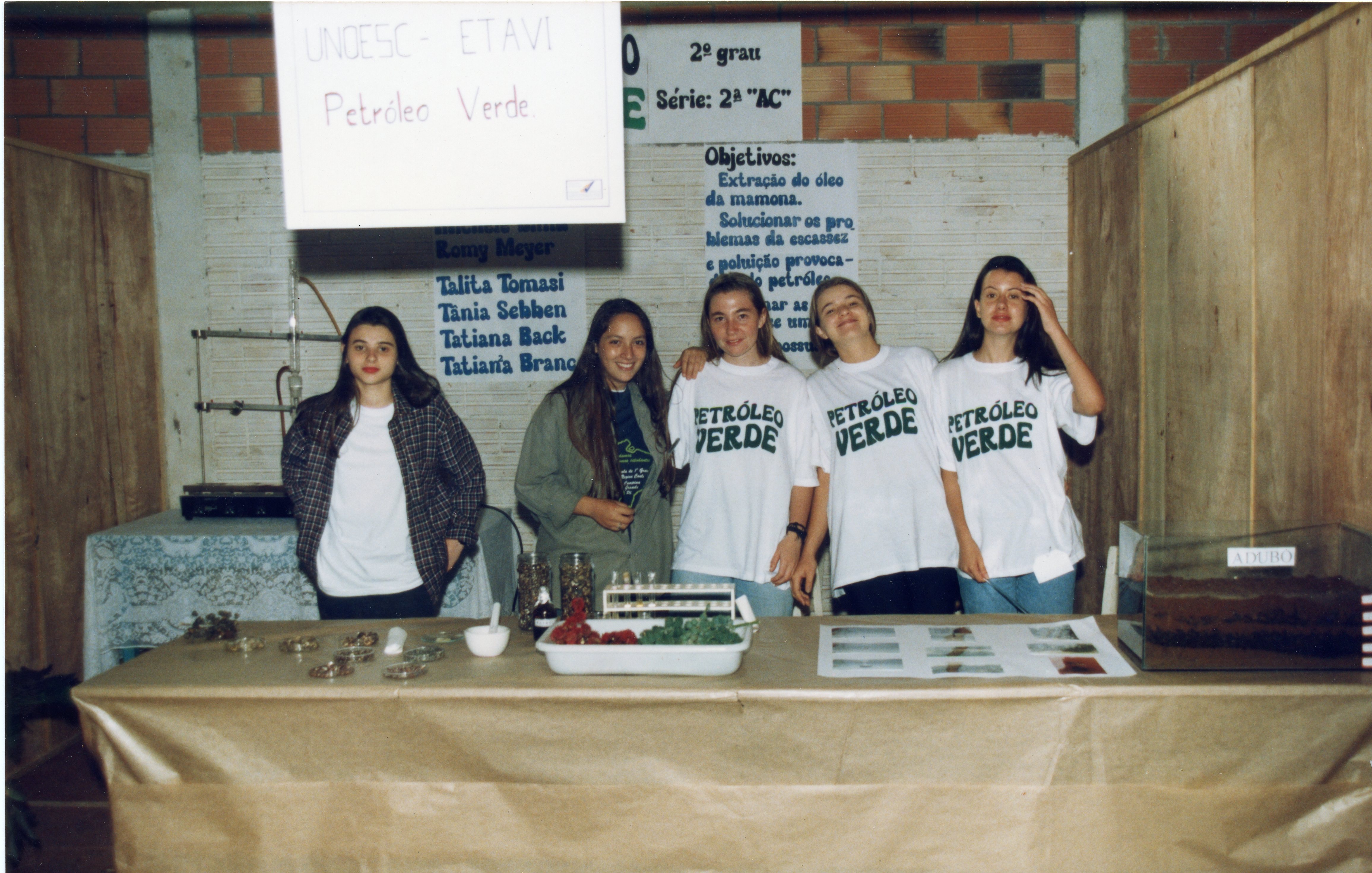 Uma fotografia de um grupo de cinco estudantes em uma feira de ciências escolar, apresentando um projeto sobre 'Petróleo Verde' (extração de óleo de mamona). O cenário inclui cartazes informativos, amostras de sementes e equipamentos de laboratório simples, sugerindo um ambiente educacional no Brasil, possivelmente na década de 1990.