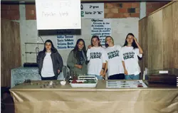 Uma fotografia de um grupo de cinco estudantes em uma feira de ciências escolar, apresentando um projeto sobre 'Petróleo Verde' (extração de óleo de mamona). O cenário inclui cartazes informativos, amostras de sementes e equipamentos de laboratório simples, sugerindo um ambiente educacional no Brasil, possivelmente na década de 1990.