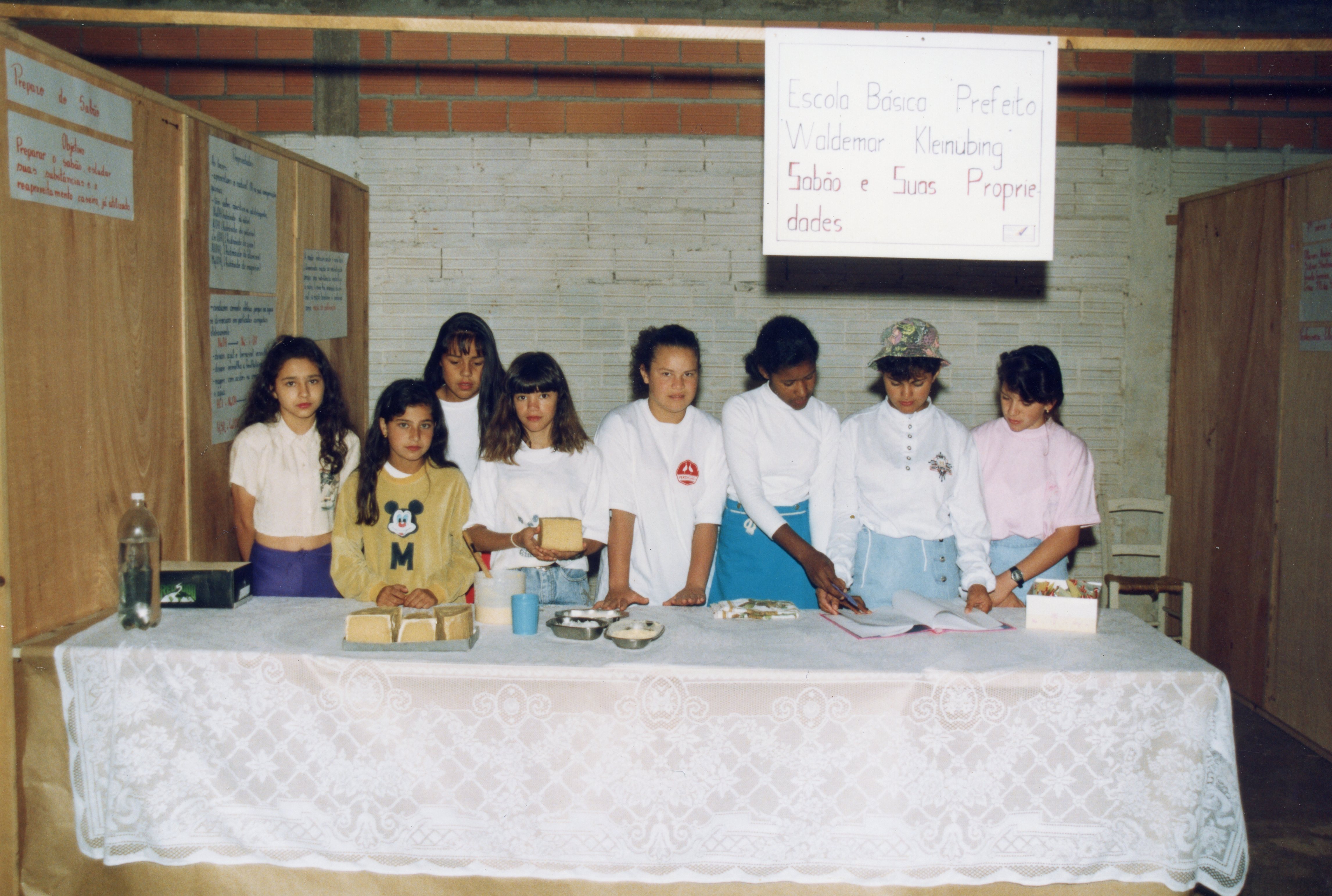 Um grupo de alunas da Escola Básica Prefeito Waldemar Kleinubing apresenta um projeto escolar sobre sabão e suas propriedades em uma feira de ciências. A cena, provavelmente da década de 1990, mostra as estudantes atrás de uma mesa coberta com toalha de renda, exibindo amostras de sabão e cartazes informativos em um ambiente escolar simples com paredes de tijolos aparentes.