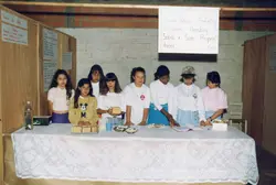 Um grupo de alunas da Escola Básica Prefeito Waldemar Kleinubing apresenta um projeto escolar sobre sabão e suas propriedades em uma feira de ciências. A cena, provavelmente da década de 1990, mostra as estudantes atrás de uma mesa coberta com toalha de renda, exibindo amostras de sabão e cartazes informativos em um ambiente escolar simples com paredes de tijolos aparentes.