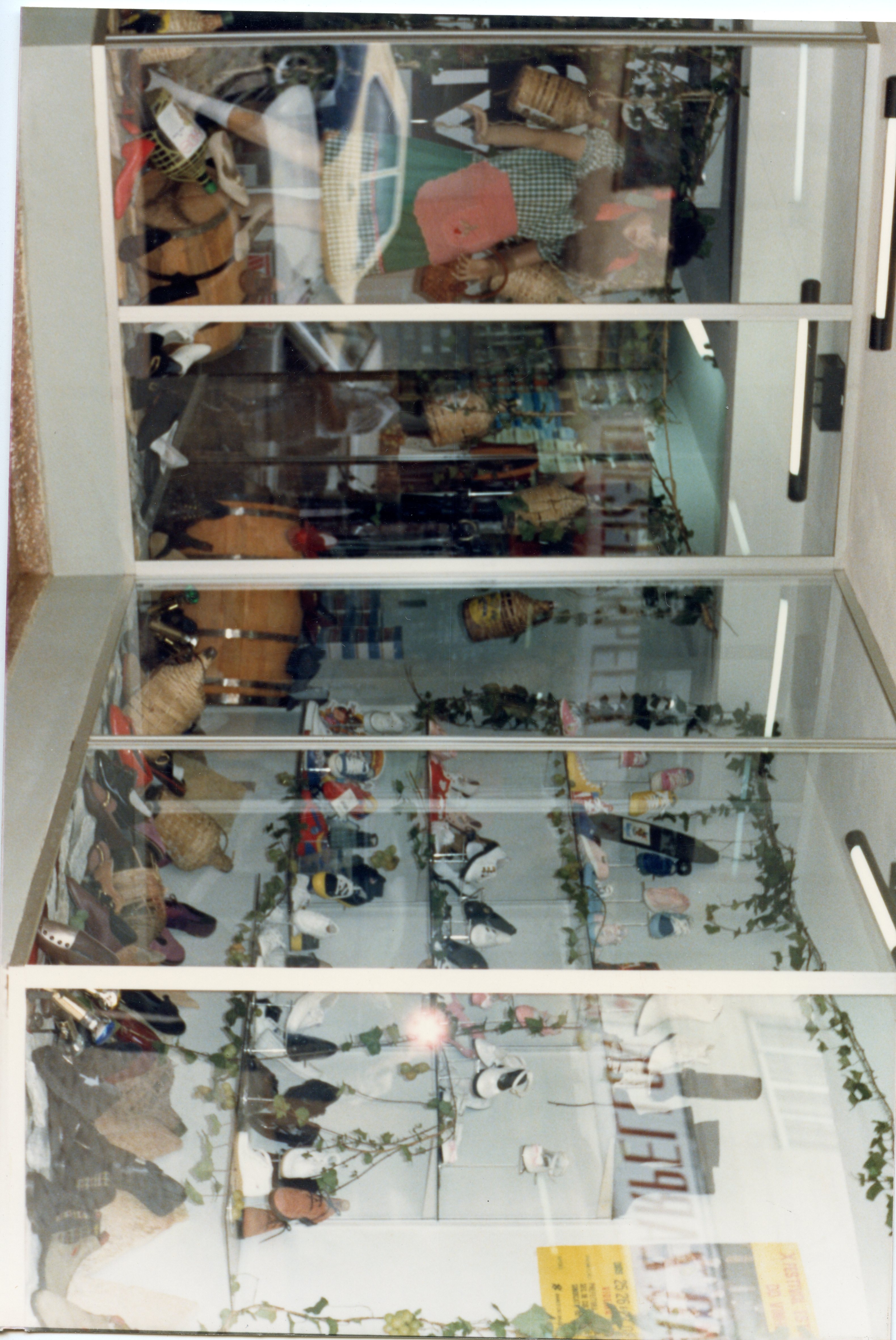 Uma fotografia de uma vitrine de loja, possivelmente uma sapataria ou comércio local, decorada com elementos rústicos como barris de madeira e cestos de vime. A imagem reflete o estilo de vitrinismo comercial de meados da década de 1990, com manequins e calçados expostos em prateleiras de vidro.