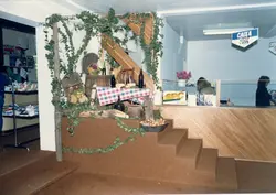 Uma fotografia interna mostrando um balcão de atendimento, possivelmente de uma cooperativa ou banco, decorado com elementos rústicos como trepadeiras, cestas de frutas e toalhas xadrez. O ambiente sugere uma celebração cultural ou temática em um espaço comercial, provavelmente nas décadas de 1980 ou 1990.