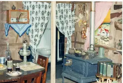 Uma cena interna que retrata um ambiente rústico, possivelmente uma cozinha ou sala de estar típica de uma casa de colonização no interior do Brasil. O espaço apresenta elementos tradicionais como um fogão a lenha, uma sanfona, um rádio antigo e utensílios domésticos, evocando uma atmosfera de nostalgia e vida rural.