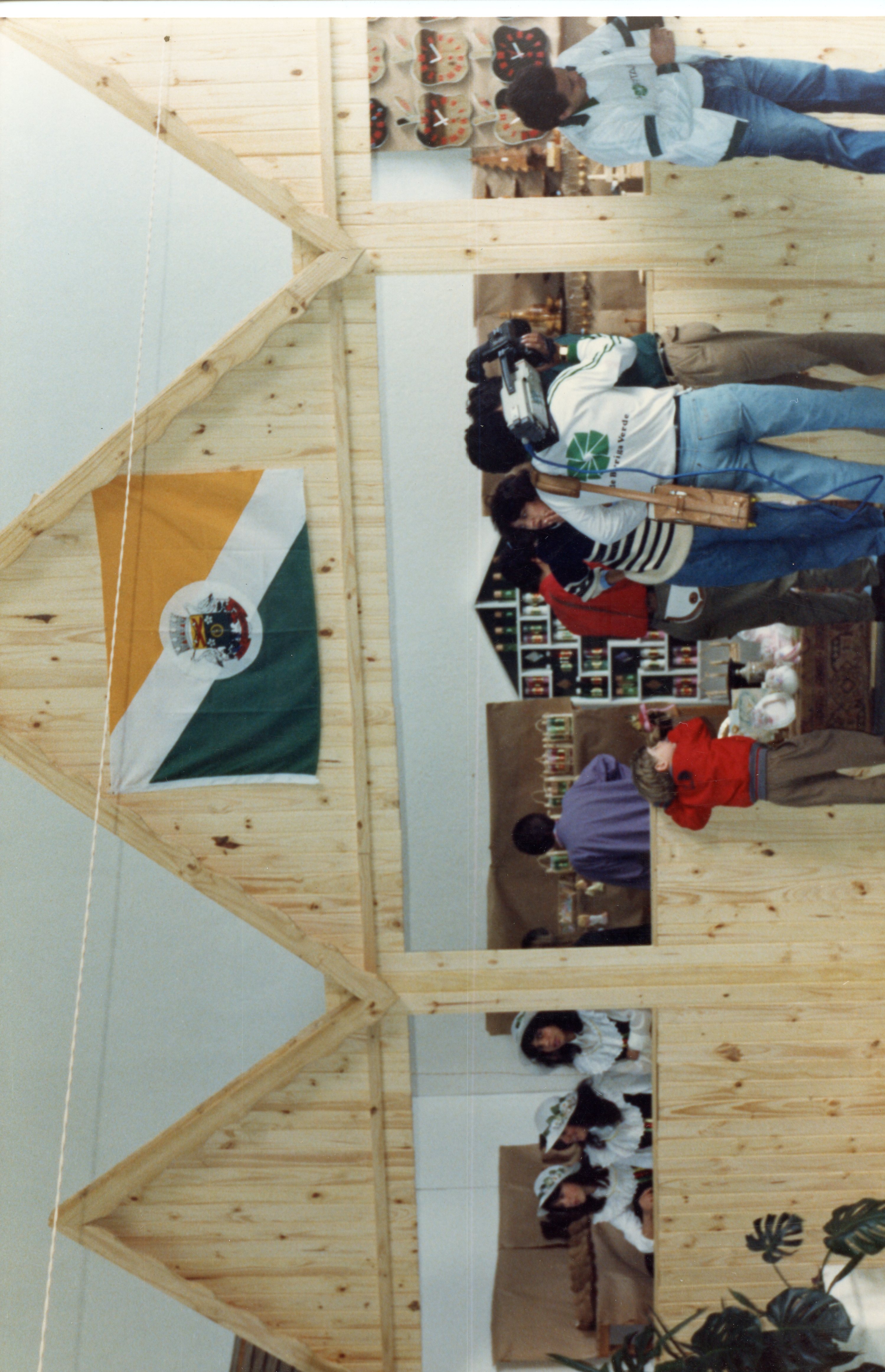 Uma fotografia que mostra um estande de madeira em uma feira ou evento, possivelmente em Videira, Santa Catarina, devido à presença da bandeira municipal. Pessoas estão presentes, incluindo um cinegrafista registrando o local, sugerindo uma cobertura jornalística ou documental de um evento regional.