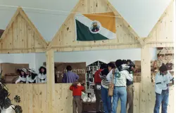Uma fotografia que mostra um estande de madeira em uma feira ou evento, possivelmente em Videira, Santa Catarina, devido à presença da bandeira municipal. Pessoas estão presentes, incluindo um cinegrafista registrando o local, sugerindo uma cobertura jornalística ou documental de um evento regional.