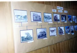 Exposição de fotografias antigas em preto e branco dispostas em uma linha do tempo sobre um grande painel de papel pardo. As imagens mostram construções e cenas urbanas, possivelmente de Videira, Santa Catarina, com legendas indicando anos e eventos históricos. A linha do tempo parece celebrar a história de uma 'Casa Centenária' ou similar.