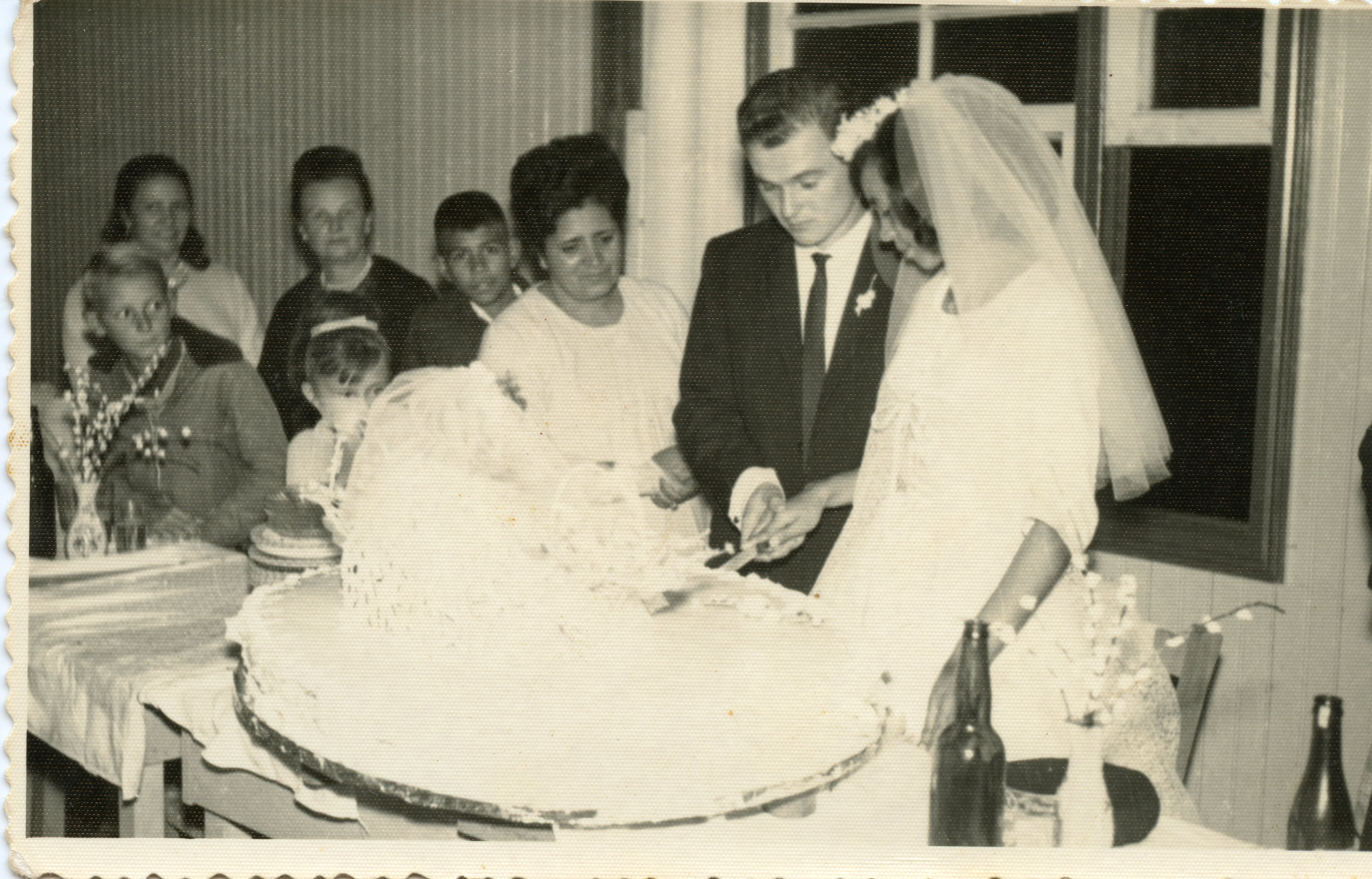Fotografia em preto e branco de um casamento, com o casal cortando o bolo. Ao fundo, familiares e convidados observam a cena. A vestimenta do casal e o estilo da fotografia sugerem uma época passada, possivelmente em meados do século XX, em um ambiente familiar e festivo.
