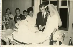 Fotografia em preto e branco de um casamento, com o casal cortando o bolo. Ao fundo, familiares e convidados observam a cena. A vestimenta do casal e o estilo da fotografia sugerem uma época passada, possivelmente em meados do século XX, em um ambiente familiar e festivo.