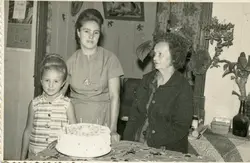 A fotografia em preto e branco mostra três mulheres em uma celebração, possivelmente um aniversário, com um bolo decorado no centro. As vestimentas e o penteado das mulheres sugerem que a foto foi tirada em meados do século XX, em um ambiente doméstico. O contexto parece ser uma reunião familiar em uma pequena cidade brasileira.