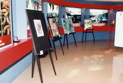 Uma exposição interna que parece ser um museu ou memorial corporativo, possivelmente da empresa Perdigão, com quadros em cavaletes e painéis fotográficos históricos nas paredes. O ambiente apresenta uma estética moderna com cores vibrantes como vermelho e azul, sugerindo uma curadoria institucional voltada para a história da indústria e desenvolvimento regional.