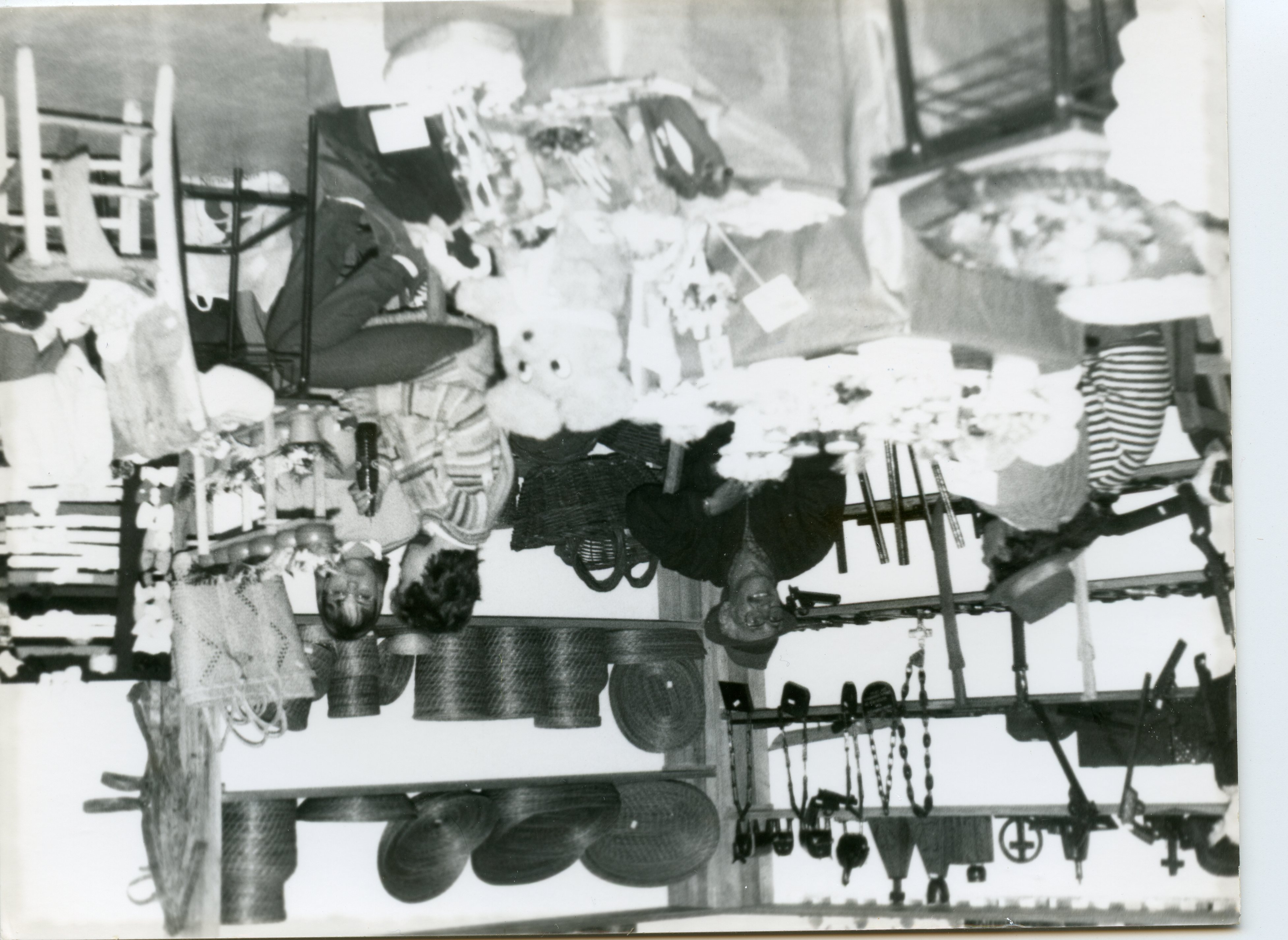 Fotografia em preto e branco que retrata um ambiente interno, possivelmente uma feira de artesanato ou comércio local, com diversas cestarias, objetos decorativos e itens de madeira expostos em prateleiras. A imagem apresenta pessoas interagindo com os produtos, sugerindo um contexto de atividade comercial ou comunitária típica de meados do século XX.