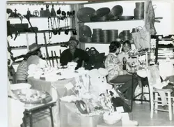 Fotografia em preto e branco que retrata um ambiente interno, possivelmente uma feira de artesanato ou comércio local, com diversas cestarias, objetos decorativos e itens de madeira expostos em prateleiras. A imagem apresenta pessoas interagindo com os produtos, sugerindo um contexto de atividade comercial ou comunitária típica de meados do século XX.