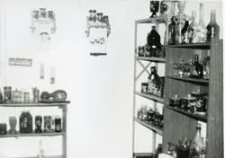 Fotografia em preto e branco exibindo prateleiras de metal carregadas com garrafas e potes decorados, possivelmente em um ambiente doméstico ou de exposição artesanal. A imagem sugere um contexto de organização ou exibição de coleções, comum em residências ou pequenos comércios de meados do século XX.