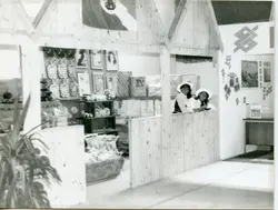 Fotografia em preto e branco de um estande de exposição, possivelmente em uma feira comercial ou evento público. Duas mulheres com chapéus estão atrás de um balcão de madeira, com produtos decorativos e o logotipo do Banco do Brasil visíveis ao fundo, sugerindo um contexto de promoção institucional ou empresarial nas décadas de 1970 ou 1980.