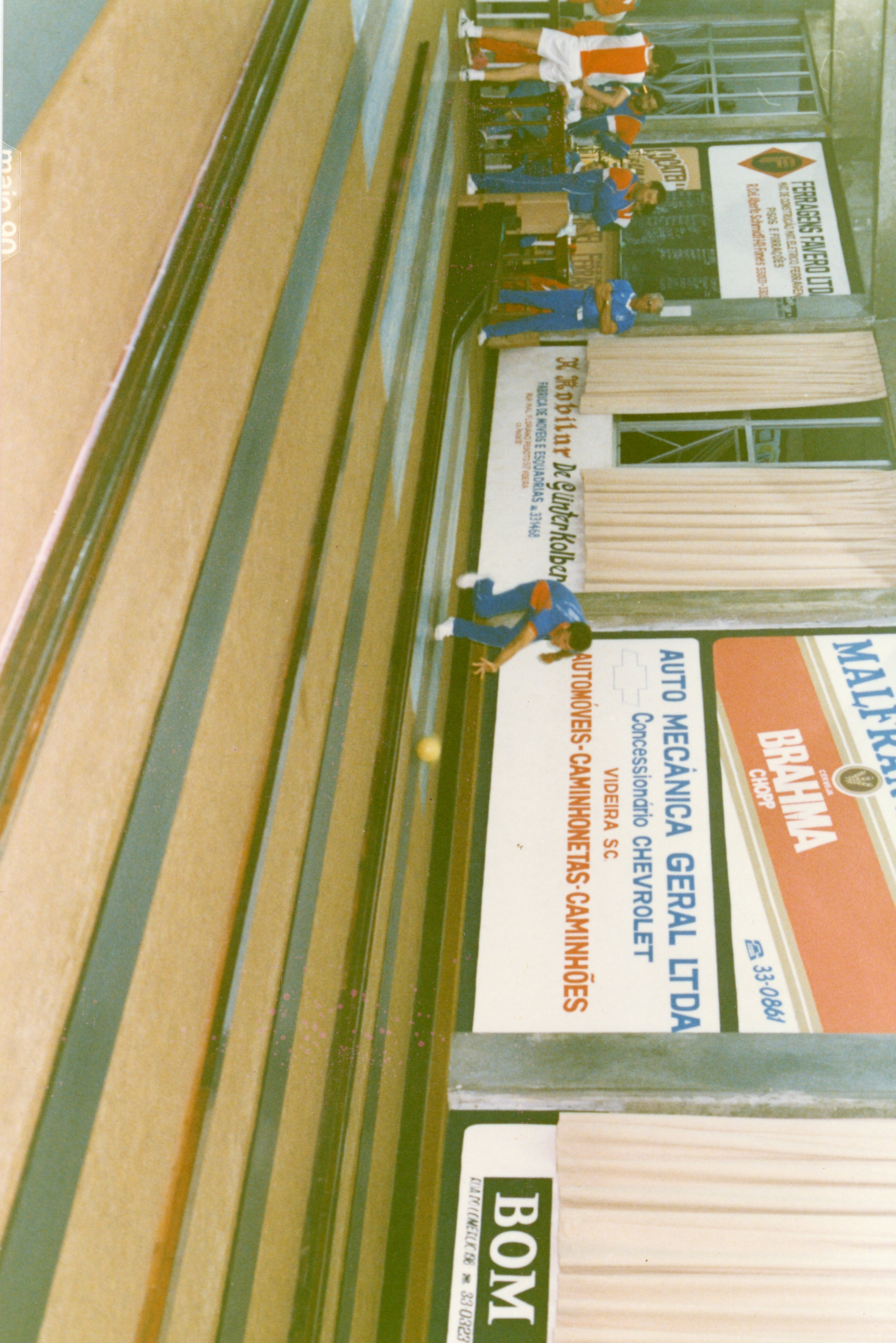 Fotografia de uma partida de bolão em um ambiente interno, provavelmente em um clube social ou esportivo em Videira, Santa Catarina. A imagem mostra jogadores em ação, com placas publicitárias de empresas locais da época visíveis nas paredes, datada de maio de 1990.