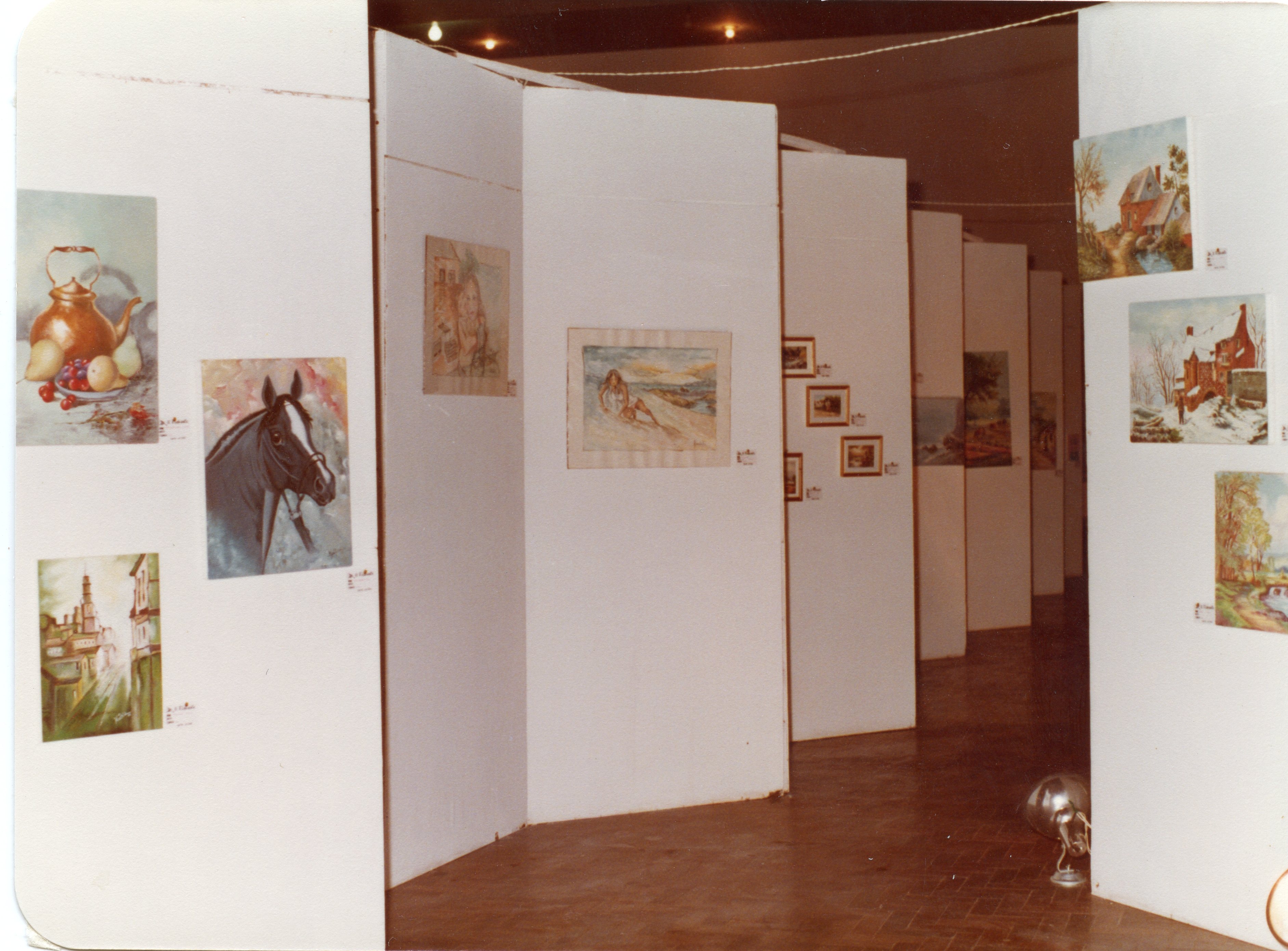 Uma exposição de arte organizada em um espaço interno com divisórias brancas dispostas em zigue-zague. As paredes exibem diversas pinturas, incluindo naturezas-mortas, retratos de animais e paisagens, sugerindo um evento cultural possivelmente ocorrido entre as décadas de 1970 e 1980.