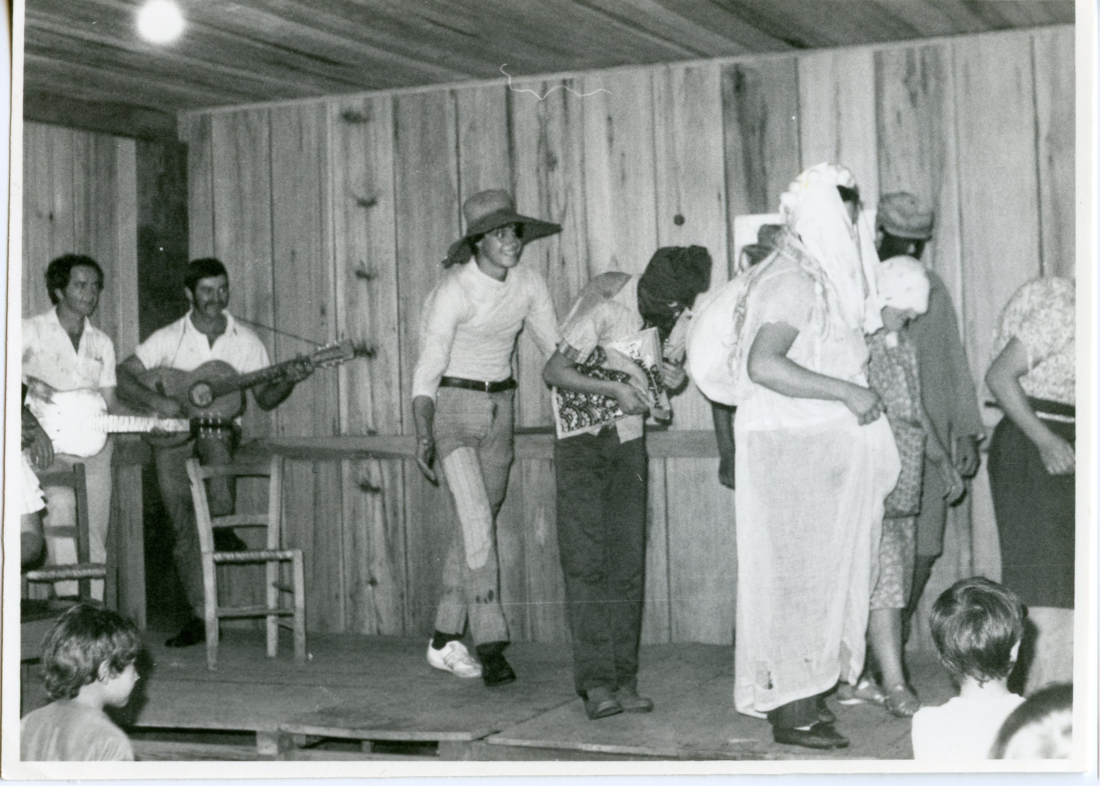 Fotografia em preto e branco registrando uma apresentação teatral ou festividade comunitária em um ambiente interno com paredes de madeira. O grupo inclui músicos com violão e pessoas fantasiadas, sugerindo uma celebração cultural ou escolar típica de pequenas cidades brasileiras em meados do século XX.