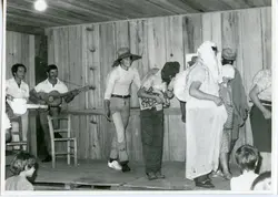 Fotografia em preto e branco registrando uma apresentação teatral ou festividade comunitária em um ambiente interno com paredes de madeira. O grupo inclui músicos com violão e pessoas fantasiadas, sugerindo uma celebração cultural ou escolar típica de pequenas cidades brasileiras em meados do século XX.