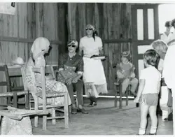 Fotografia em preto e branco retratando um ambiente interno rústico, possivelmente uma casa de madeira ou galpão, com várias pessoas reunidas. Mulheres vestidas com aventais e lenços na cabeça interagem enquanto uma criança observa a cena, sugerindo um contexto de vida comunitária ou rural, possivelmente em meados do século XX.