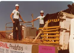 Fotografia colorida retratando dois jovens em um caminhão de transporte de animais, provavelmente em uma exposição agropecuária ou evento rural. Eles vestem camisetas com o emblema do Clube 4-S, uma organização voltada para a juventude rural, e acompanham bovinos em uma estrutura de madeira, sugerindo uma atividade de demonstração ou feira agrícola típica de cidades do interior brasileiro.