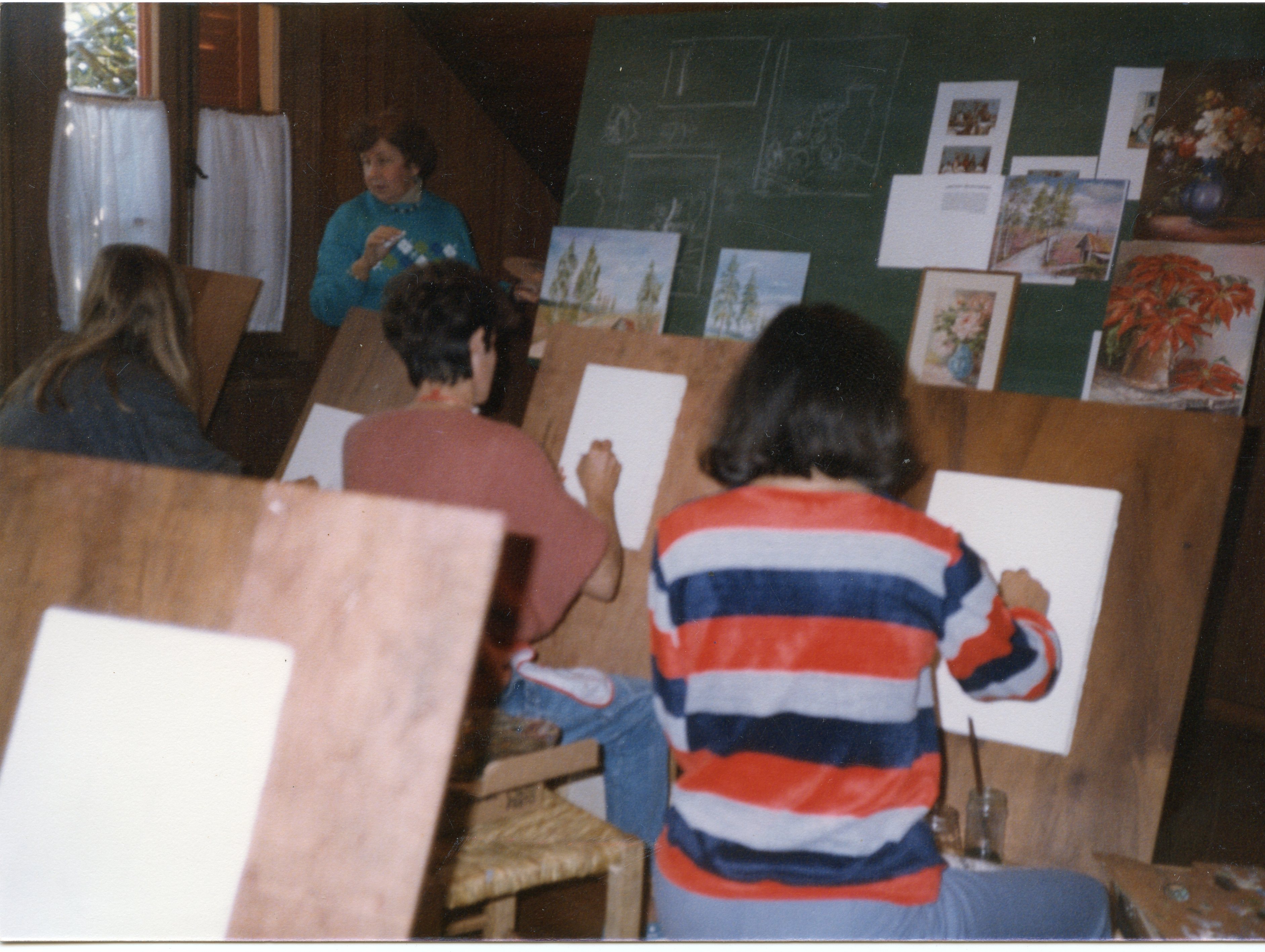 Uma aula de pintura em um ambiente interno, possivelmente um ateliê, onde alunos trabalham em cavaletes de madeira. A instrutora observa os alunos enquanto quadros de referência estão fixados em um quadro negro ao fundo, sugerindo uma atividade educacional artística típica das décadas de 1980 ou 1990.