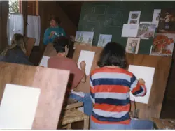 Uma aula de pintura em um ambiente interno, possivelmente um ateliê, onde alunos trabalham em cavaletes de madeira. A instrutora observa os alunos enquanto quadros de referência estão fixados em um quadro negro ao fundo, sugerindo uma atividade educacional artística típica das décadas de 1980 ou 1990.