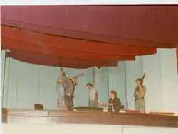 Uma fotografia colorida que registra uma encenação teatral ou apresentação em um palco, com cinco pessoas vestidas com roupas simples e chapéus, portando o que parecem ser espingardas cenográficas. O cenário é composto por cortinas vermelhas e painéis claros, sugerindo uma produção cultural ou escolar típica de meados da década de 1970 ou 1980.