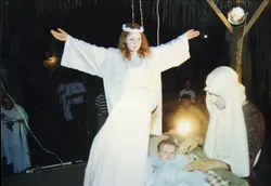 Uma encenação teatral de um presépio vivo, provavelmente realizada em um contexto comunitário ou religioso. A imagem mostra pessoas vestidas como personagens bíblicos, incluindo um anjo com braços abertos e uma representação da Sagrada Família com um bebê, sob iluminação noturna.